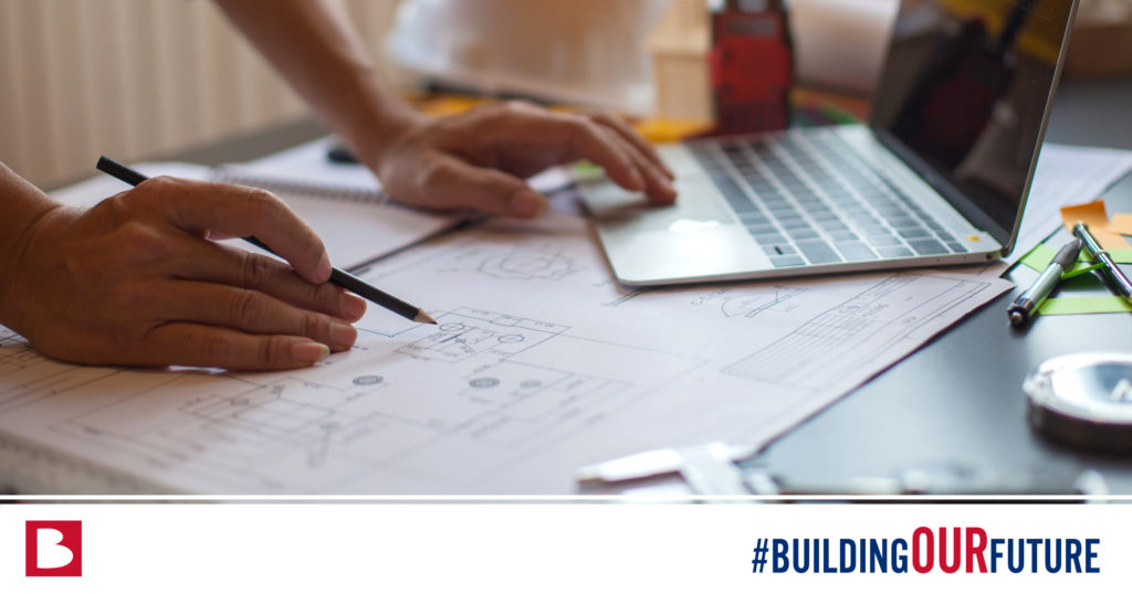 person leaning over a desk containing a laptop and floor plans with the hashtag #buildingourfuture